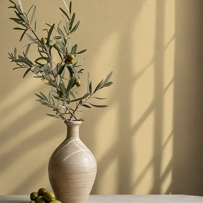 Glööckler Pompöös Provence Vliestapete - Gelb, Uni, 10,05 x 0,53 m - Nahaufnahme einer beigen Vase mit einem Olivenzweig und Oliven auf einem Tisch vor einer schlichten, beigen Wand.