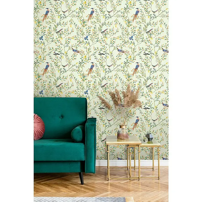 Glööckler Pompöös Provence Vliestapete - Grün/Orange/Gelb, Floral, 10,05 x 0,53 m - Grünes Samtsofa mit rosa Kissen, goldenen Beistelltischen und einer Vase mit Pampasgras vor einer Tapete mit Vogel- und Zitronenmuster.