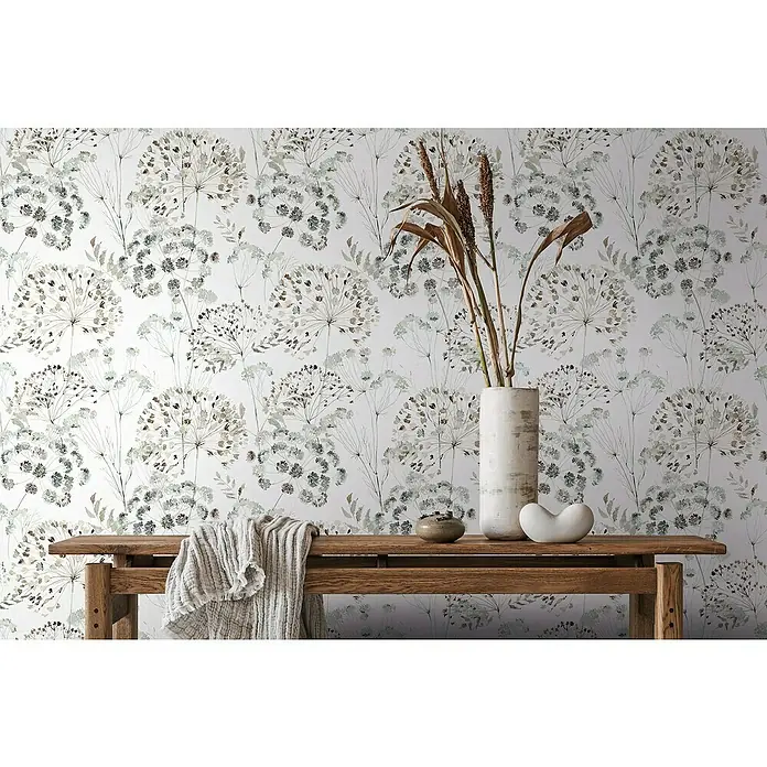 Glööckler Pompöös Provence Vliestapete - Grau/Beige/Grün, Floral, 10,05 x 0,53 m - Das Bild zeigt eine Wand mit einem floralen Tapetenmuster, einem Holztisch und dekorativen Objekten wie einer Vase und einer Keramikschale.