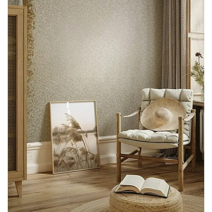 Glööckler Pompöös Provence Vliestapete Basic - Beige, Floral, 10,05 x 0,7 m - Ein gemütliches Interieur mit einem bequemen Sessel, dekoriert mit einem Strohhut, und einem offenen Buch auf einem Hocker.