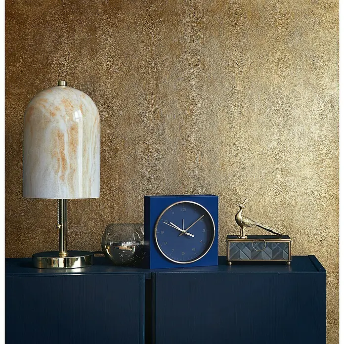 Glööckler Pompöös Provence Vliestapete - Gold, Metalloptik, 10,05 x 0,53 m - Nahaufnahme einer blauen Uhr, einer Lampe und einer dekorativen Vogelstatue auf einem dunklen Schrank vor goldenem Hintergrund.