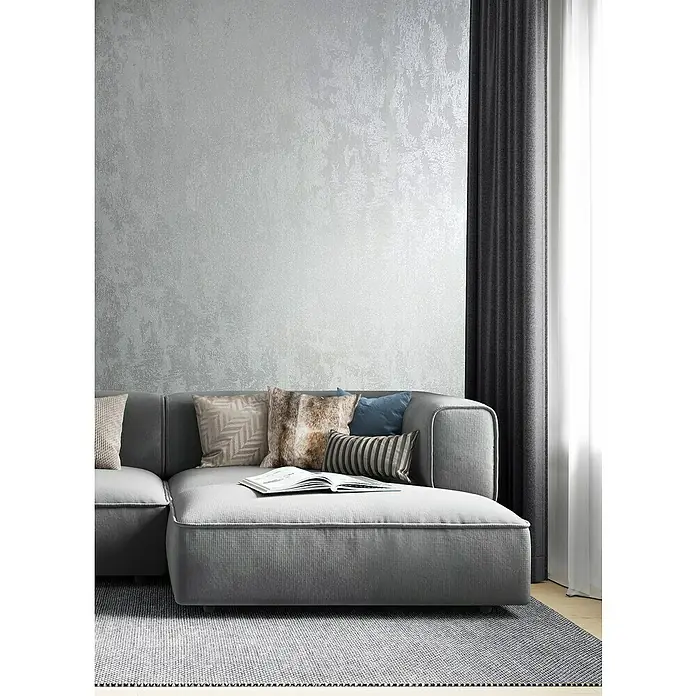 Glööckler Pompöös Provence Vliestapete - Silber, Metalloptik, 10,05 x 0,53 m - Elegantes graues Sofa mit Zierkissen und einem offenen Buch. Das Sofa steht auf einem grauen Teppich vor einer silbernen Wand.