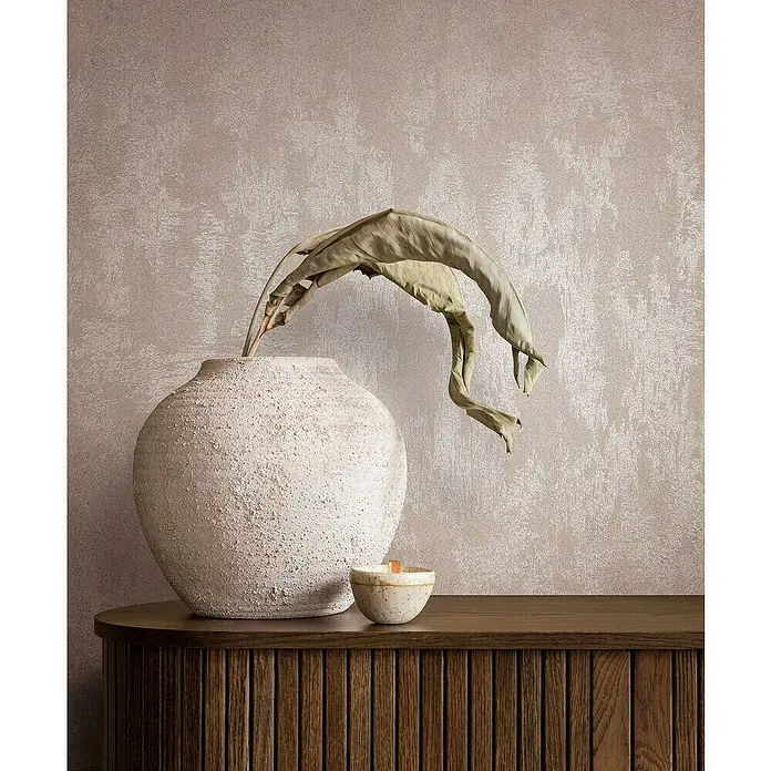 Glööckler Pompöös Provence Vliestapete - Rosa, Metalloptik, 10,05 x 0,53 m - Dekorative Szene mit einer strukturierten Vase, einer Kerze und einem getrockneten Blatt auf einem Holzschrank vor einer strukturierten Wand.