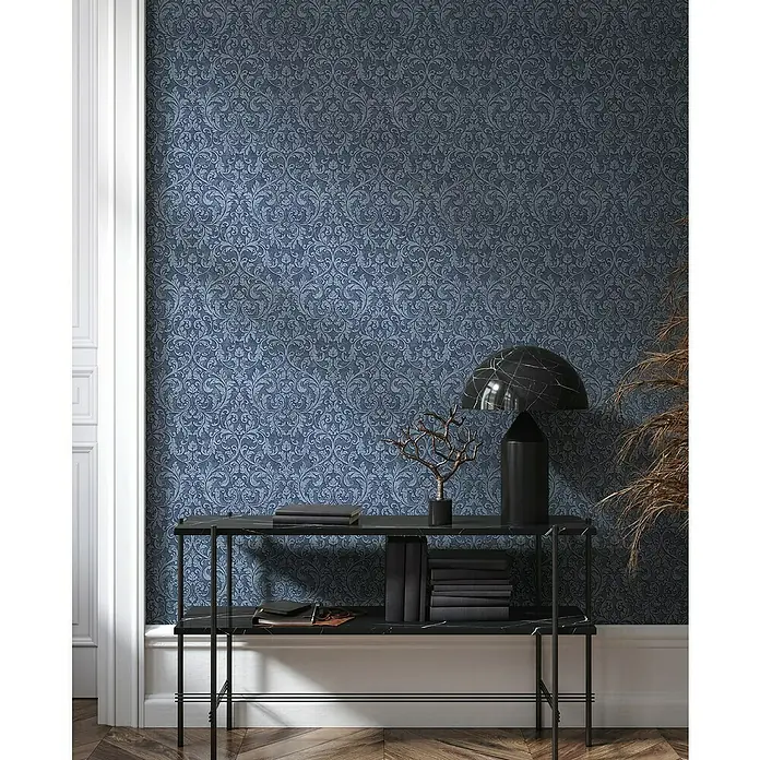 Glööckler Pompöös Provence Vliestapete - Blau/Schwarz, Ornament, 10,05 x 0,53 m - Elegante Konsole mit Marmorplatte und Bücherstapel vor einer Wand mit blauem, floralem Muster.