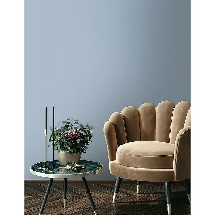 Glööckler Pompöös Provence Vliestapete - Blau, Uni, 10,05 x 0,53 m - Nahaufnahme eines beigefarbenen Muschelsessel neben einem Marmortisch mit Kerzenständern und Blumenvase vor einer blauen Wand.