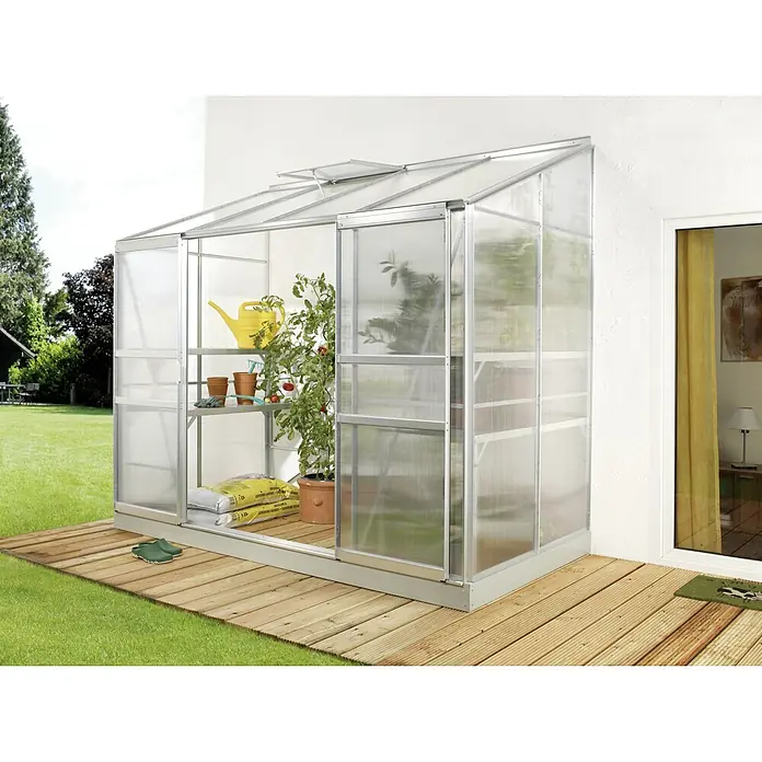 Vitavia Gewächshaus Styx 3300 - Außenmaß inkl. Dachüberstand (B x T): 255 x 132 cm, 4 mm, Aluminium - Ein Gewächshaus aus Aluminium und Polycarbonat steht auf einer Holzterrasse, gefüllt mit Pflanzen und Gartenzubehör.