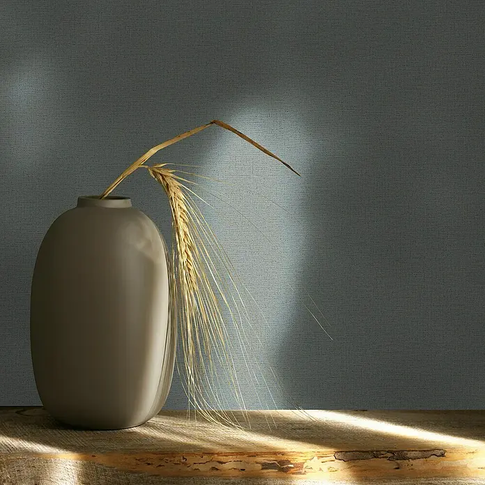 Glööckler Pompöös Provence Vliestapete - Mittelgrau, Uni, 10,05 x 0,53 m - Nahaufnahme einer grauen Keramikvase mit goldenen Weizenähren auf einem Holztisch. Das sanfte Licht betont die Texturen.