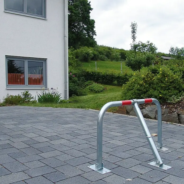 Schellenberg Parkbügel - 440 x 820 x 560 mm, Stahl - Eine silberne Fahrradanlehnbügel mit roten Griffen steht auf einem gepflasterten Untergrund vor einem Haus und Grünflächen.