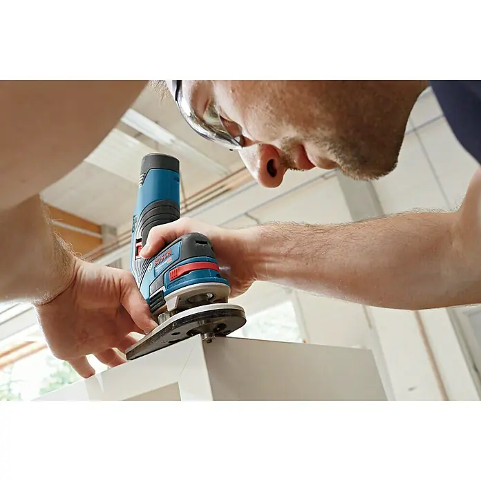 Bosch Professional Akku-Kombifräse GKF 12V-8 L-Boxx - 12 V, Ohne Akku, Leerlaufdrehzahl: 13.000 U/min - Nahaufnahme einer Bosch Akku-Kantenfräse beim Bearbeiten einer weißen Holzoberfläche, bedient von einer Person mit Schutzbrille.