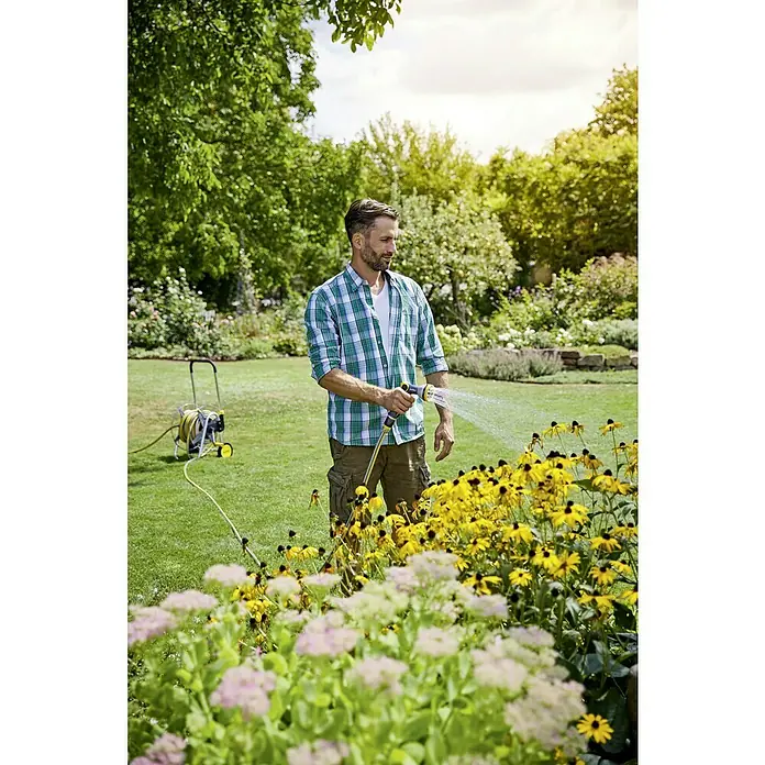 Neptun Profi Pistola multiuso - Aluminio/Plástico - Un hombre riega flores en su jardín con una manguera de jardín amarilla. El césped es verde y bien cuidado.