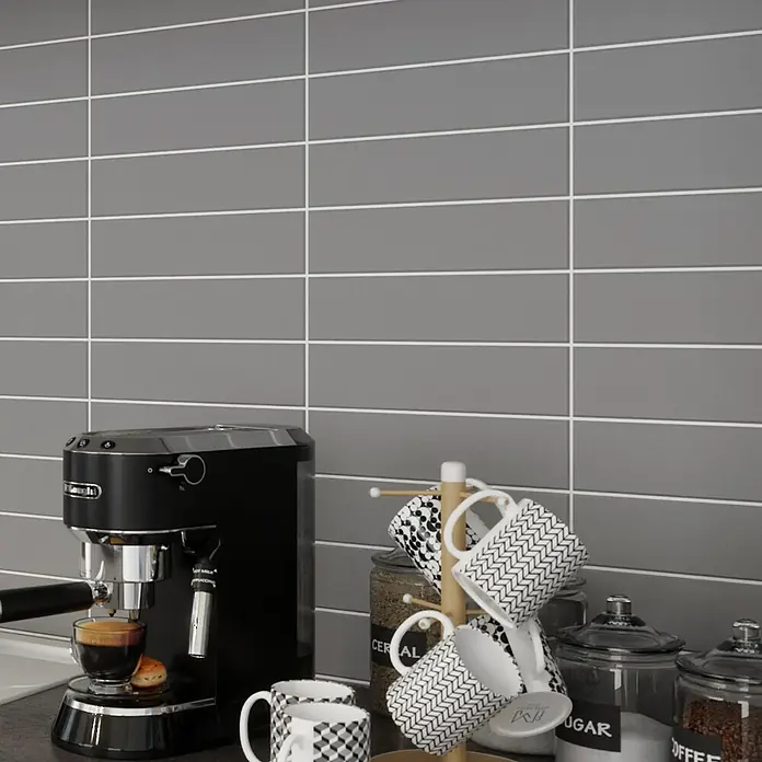 Modern Style Wandfliese Urban Gris - 7,5 x 30 cm, Grau, Matt - Nahaufnahme einer Delonghi Espressomaschine mit Tassen, einem Tassenbaum und Vorratsgläsern auf einer grauen Arbeitsplatte vor grau gefliestem Hintergrund.