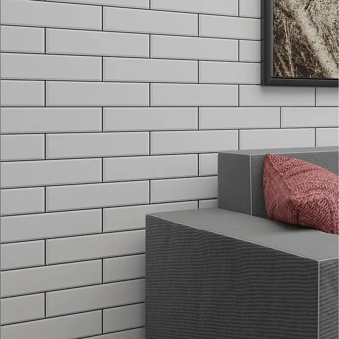 Modern Style Wandfliese Manhattan Biselado Blanco - 7,5 x 30 cm, Weiß, Matt - Ein Bild von länglichen weißen Fliesen mit dunklen Fugen an einer Wand hinter einem grauen Sofa mit einem roten Kissen.