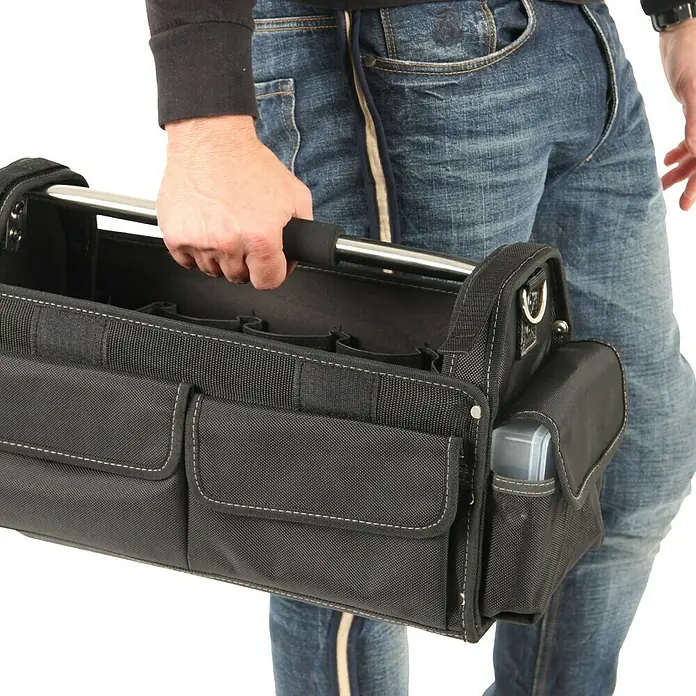 Hepco & Becker Werkzeugtasche Profi-Installateurtasche 5858 - L x B x H: 200 x 400 x 240 mm, Polyester - Eine schwarze Werkzeugtasche mit silbernem Griff wird von einer Person in Jeans gehalten. Die Tasche hat mehrere Fächer und weiße Nähte.