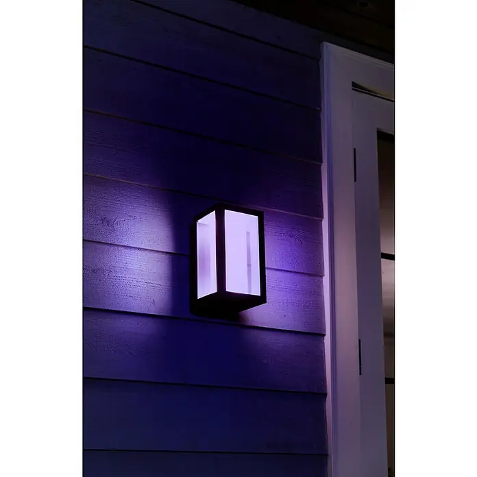Philips Hue LED-Außenwandleuchte Impress - 8 W, 14,1 x 12 x 24 cm, Schwarz, IP44 - Moderne Außenleuchte mit violettem Licht an einer blauen Holzwand neben einer weißen Tür. Die Leuchte ist rechteckig und hat ein minimalistisches Design.