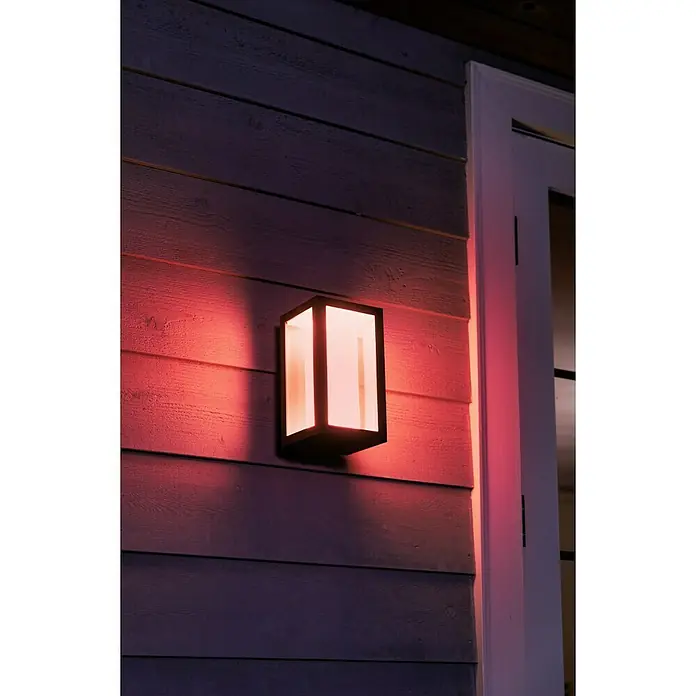 Philips Hue LED-Außenwandleuchte Impress - 8 W, 14,1 x 12 x 24 cm, Schwarz, IP44 - Eine moderne Außenwandleuchte mit rotem Licht illuminiert eine Hauswand und einen weißen Türrahmen und sorgt so für eine angenehme Atmosphäre.