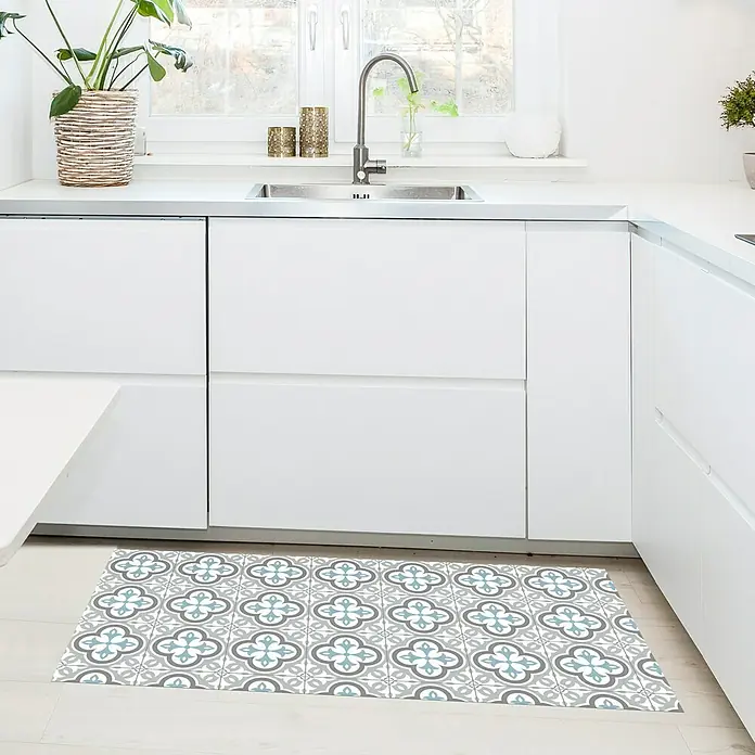 Alfombra de vinilo Croma Hidra topo - 110 x 50 cm - Una alfombra de cocina decorativa con un patrón floral azul grisáceo se encuentra frente a una moderna cocina blanca con fregadero y grifo.