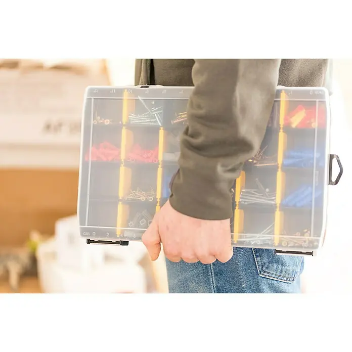 Stanley Estuche para piezas pequeñas STST81681-1 - Número de compartimentos: 22 ud., Sin contenido, Negro/Amarillo - Una persona lleva una caja de surtido transparente llena de tornillos y tacos de varios colores y tamaños. Ideal para talleres y proyectos de bricolaje.