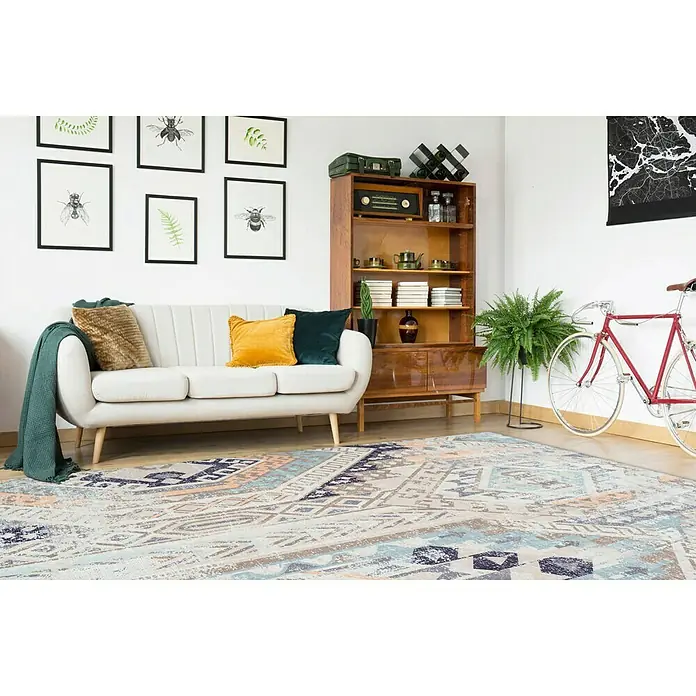 Kayoom Kurzflorteppich IndianaMulti/Blau, 230 x 160 cm, 100 % Baumwolle - Ein stilvoller Raum mit einem bunten Teppich mit geometrischen Mustern im Vordergrund, ergänzt durch Möbel wie Sofa und Bücherregal.