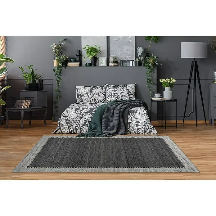 Kayoom Flachgewebeteppich Phoenix - Anthrazit/Grau, 150 x 80 cm, 75 % Wolle - Hier sehen wir einen grauen Teppich mit dekorativem Rand in einem Schlafzimmer, der dem Raum einen Hauch von Eleganz verleiht.