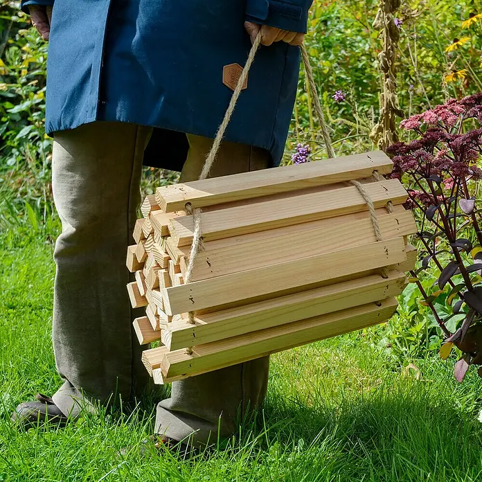 Dobar Gartenweg Mobil - L x B x H: 195 x 36 x 2 cm, Transportfähig - Nahaufnahme einer Person, die ein Holzscheitregal mit Seilgriffen trägt, umgeben von Rasen und Pflanzen. Das Holzregal ist der Schwerpunkt des Bildes.