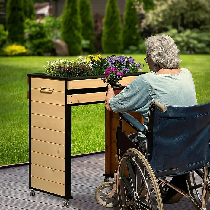 Dobar Hochbeet Wheely auf Rollen - 77 x 47 x 88 cm, Holz, Braun/Schwarz - Eine ältere Frau im Rollstuhl gärtnert an einem rollbaren, höhenverstellbaren Pflanztisch mit Blumen bepflanzt, der Gartenarbeit zugänglicher macht.