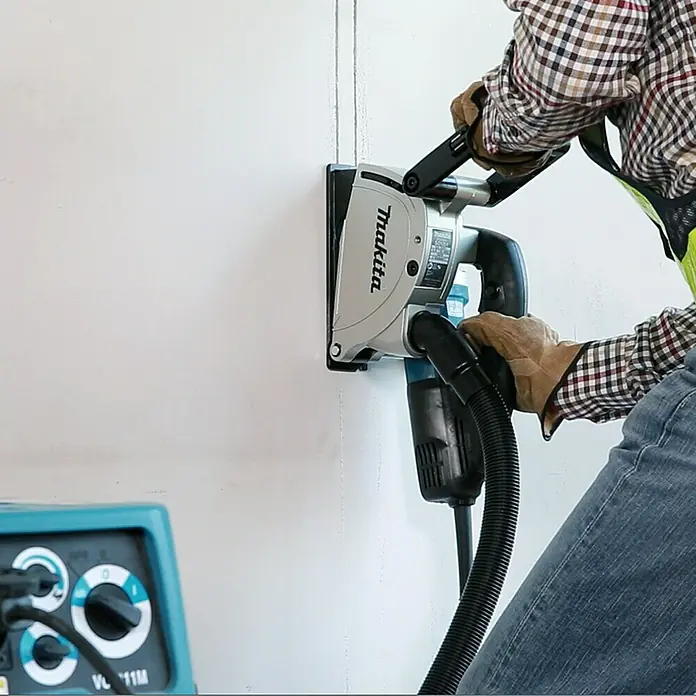 Makita Mauernutfräse SG1251J - 1.400 W, Durchmesser Scheibe: 125 mm - Ein Arbeiter benutzt eine Makita Mauernutfräse an einer weißen Wand, um präzise Schlitze zu erstellen. Das Gerät ist mit einem Staubsauger verbunden.