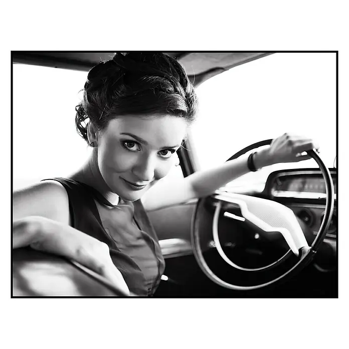 Cuadro Girl - Chica al volante, An x Al: 40 x 30 cm - Foto en blanco y negro de una mujer en un coche antiguo, sonriendo con confianza y sujetando el volante.