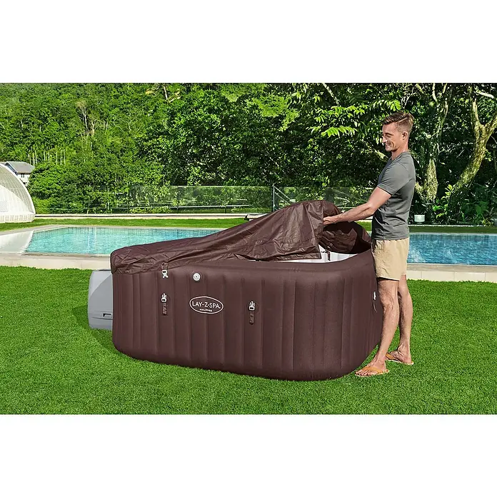 Bestway Lay-Z-Spa Mobil-Whirlpool Maldives HydroJet - L x B x H: 201 x 201 x 80 cm, Mahagoni, 1.050 l - Ein Mann öffnet den braunen Lay-Z-Spa Whirlpool, der auf einem grünen Rasen neben einem Pool steht.