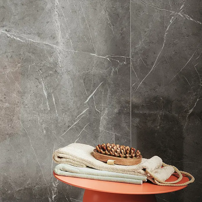 Atlas Concorde Solution Marble Vison Feinsteinzeugfliese Grigio Chiaro - 60 x 120 cm, Grau, Glänzend - Das Bild zeigt eine orangefarbene Badezimmerablage mit Handtüchern und einer Massagebürste vor einer grauen Marmorwand.