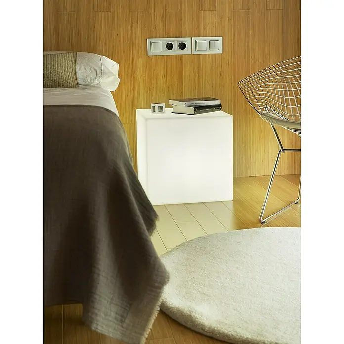 New Garden Lámpara LED decorativa Cuby 40 - Blanco, L x An x Al: 40 x 40 x 40 cm, 650 lm - Una mesita auxiliar cúbica blanca iluminada junto a un lecho. Contiene libros y un reloj digital, ofreciendo luz y superficie.