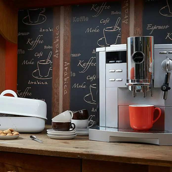 A.S. Création Designpanel pop.up - Espresso Arabica, Schwarz/Braun, 35 cm x 2,5 m - Nahaufnahme einer Jura-Kaffeemaschine mit Tassen und Keksen auf einem Holztisch vor einer Kaffeetapete.