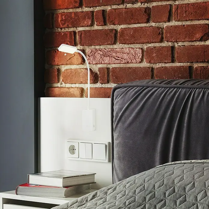 Briloner LED-Leseleuchte Cielo - Höhe: 56 cm, Weiß - Nahaufnahme einer eleganten Nachttischlampe mit flexiblem Arm, montiert auf einem weißen Paneel mit Steckdose und Schaltern, vor einer Backsteinwand.