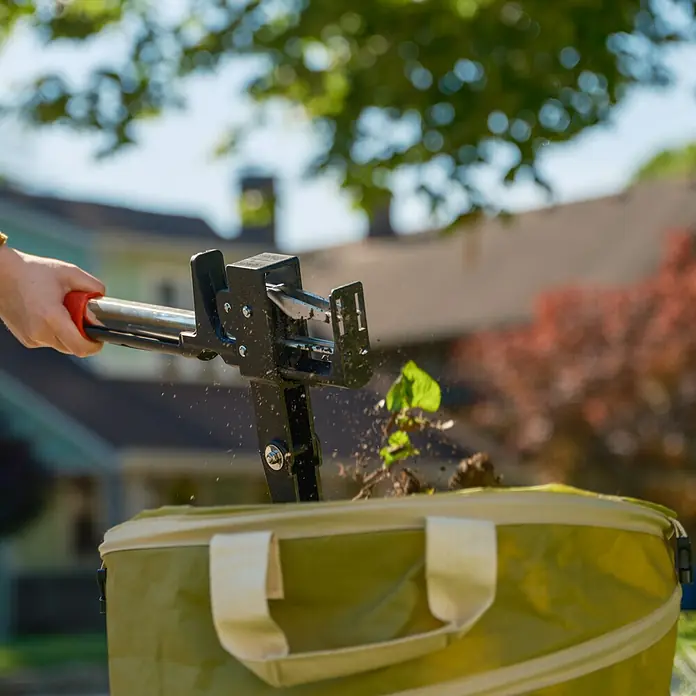 Root Slayer Unkrautstecher - Länge: 100 cm - Nahaufnahme eines Unkrautstechers, der Unkraut über einem grünen Gartenabfallsack auswirft, mit verschwommenem Hintergrund.
