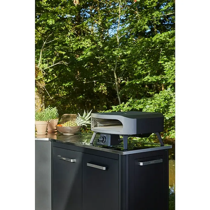 Cozze Pizzaofen Classic 17" - Grillfläche (B x T): 42,5 x 42,5 cm, 8 kW - Nahaufnahme eines Cozze Pizzaofens im Freien mit Edelstahl-Arbeitsplatte, Schränken, Töpfen und Obst.