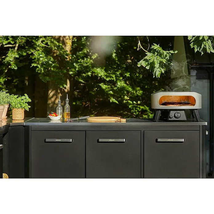 Cozze Pizzaofen Classic 13 - Grillfläche (B x T): 34 x 34 cm, 6 kW - Eine dunkelgraue Outdoor-Küchenzeile mit einem silbernen Pizzaofen, der eine Pizza backt, umgeben von frischen Kräutern und Gewürzen auf der Arbeitsplatte.