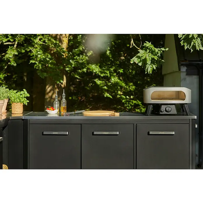 Cozze Pizzaofen Classic 13 - Grillfläche (B x T): 34 x 34 cm, 6 kW - Dunkelgraue Outdoor-Küche mit silbernem Pizzaofen auf der Arbeitsplatte, umgeben von frischen Kräutern und Kochutensilien.