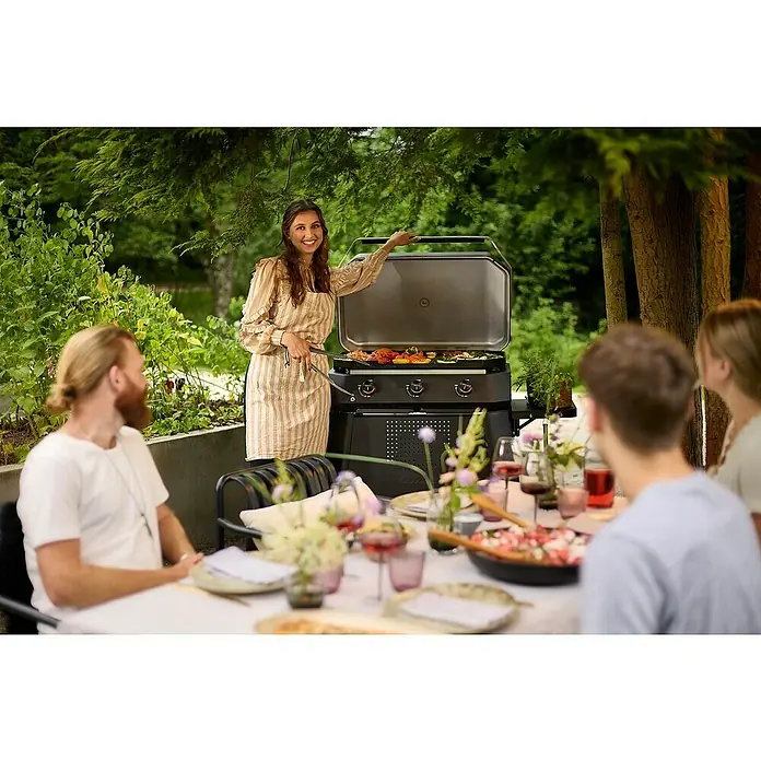 Cozze Plancha-Grill 800 - Grillfläche (B x T): 80 x 50 cm, 7,5 kW, Schwarz - Eine Frau grillt Gemüse auf einem Gozney Grill bei einer Gartenparty. Der Grill steht im Mittelpunkt einer geselligen Runde.