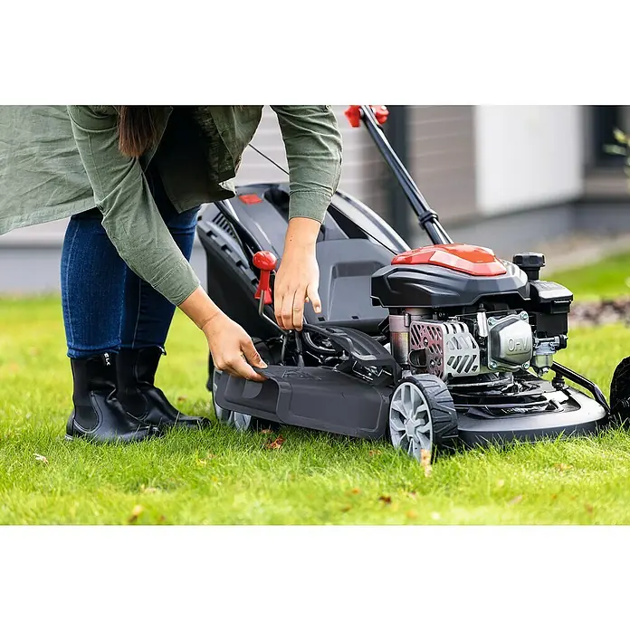 Scheppach Benzin-Rasenmäher PML46-150ES - 150 cm³, Schnittbreite: 46 cm, Hybrid-Start: Elektrostart & Seilzugstarter - Eine Frau bedient einen Rasenmäher im Freien. Der Rasenmäher ist rot und schwarz. Die Frau trägt blaue Jeans und schwarze Stiefel.