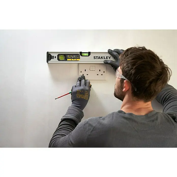 Stanley FatMax Nivel de burbuja Xtreme - Plateado - Un hombre usa un nivel Stanley para alinear un enchufe eléctrico en una pared blanca, con gafas de seguridad y guantes.
