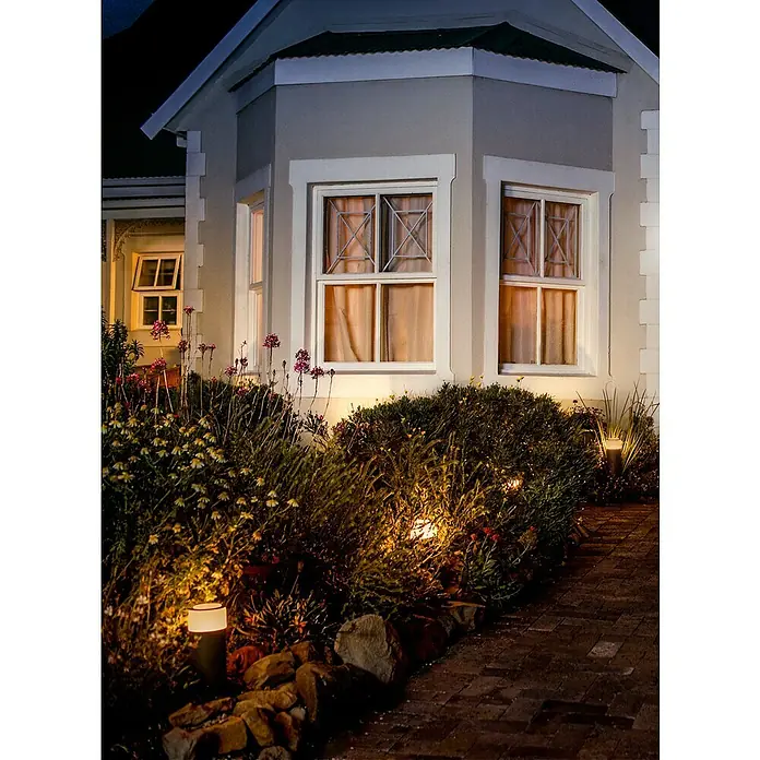 Philips Hue Außensockelleuchte Calla Erweiterung - 8 W, Farbe Gehäuse: Schwarz, RGBW, 10,4 x 10,4 x 25,2 cm, Mit Leuchtmittel - Das Bild zeigt eine Hausfassade mit beleuchteten Fenstern und Garten. Die Außenbeleuchtung setzt Akzente und betont die Architektur des Hauses.