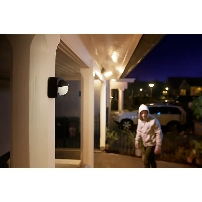 Philips Hue Bewegungsmelder - Reichweite Sensor: 12 m, Schwarz, L x B x H: 7,5 x 17,6 x 16,5 cm - Eine moderne Bewegungsmelder-Außenleuchte mit einem verdächtigen Mann im Hintergrund, die Sicherheit und Komfort vereint.