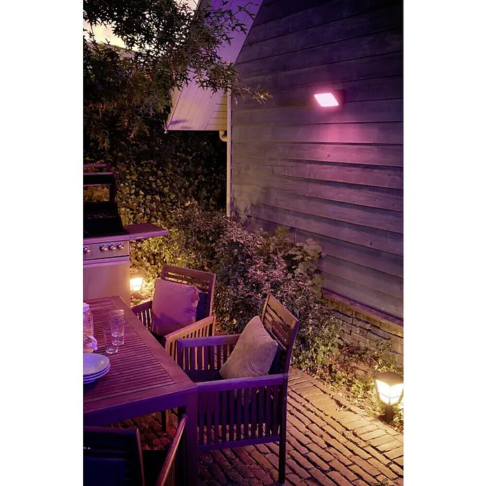 Philips Hue LED-Strahler White & Color Ambiance - 15 W, L x B x H: 22 x 16 x 15,3 cm, Schwarz, RGBW - Beleuchteter Gartenbereich mit stilvollen Gartenmöbeln und Grill, stimmungsvoll in Szene gesetzt durch farbige Außenbeleuchtung.
