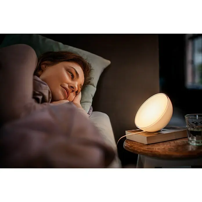 Philips Hue LED-Tischleuchte Go - 6 W, L x B x H: 15 x 15 x 7,9 cm, RGBW - Frau im Bett mit einer Philips Hue Lampe auf dem Nachttisch, die sanftes Licht spendet und eine entspannende Atmosphäre schafft.