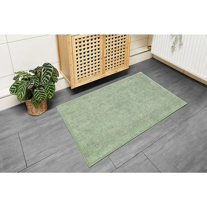 Badteppich Malibu - 60 x 100 cm, Salbei, 100% Polyester - Ein hellgrüner Badteppich liegt auf einem grauen Fliesenboden neben einem Bambusschrank und einer Pflanze.