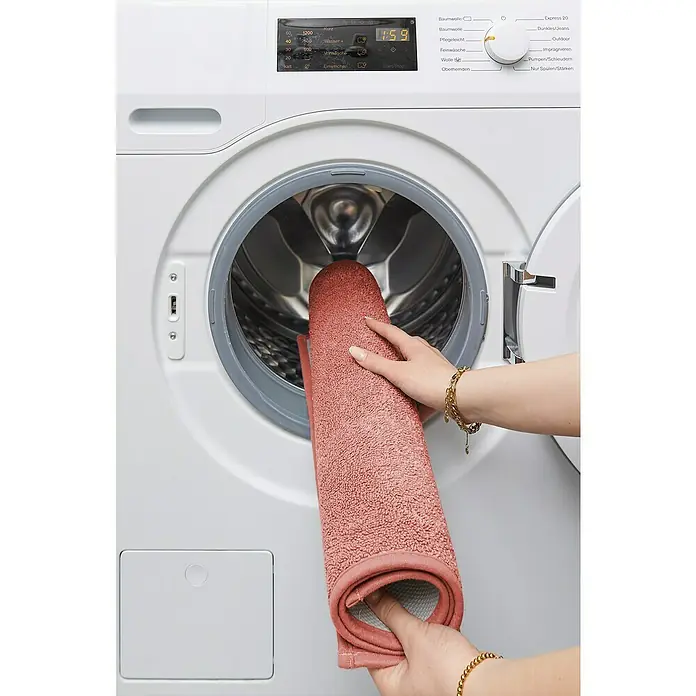 Badteppich Malibu - 60 x 100 cm, Rostrot, 100% Polyester - Eine Person legt eine rosafarbene Badematte in eine weiße Waschmaschine.