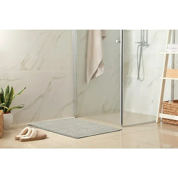 Badteppich Atlanta - 50 x 80 cm, Grau, 100% Baumwolle - Ein Badezimmer-Setup mit einer hellgrauen Badematte vor einer Duschkabine, dazu Hausschuhe, eine Pflanze und ein geflochtener Korb.