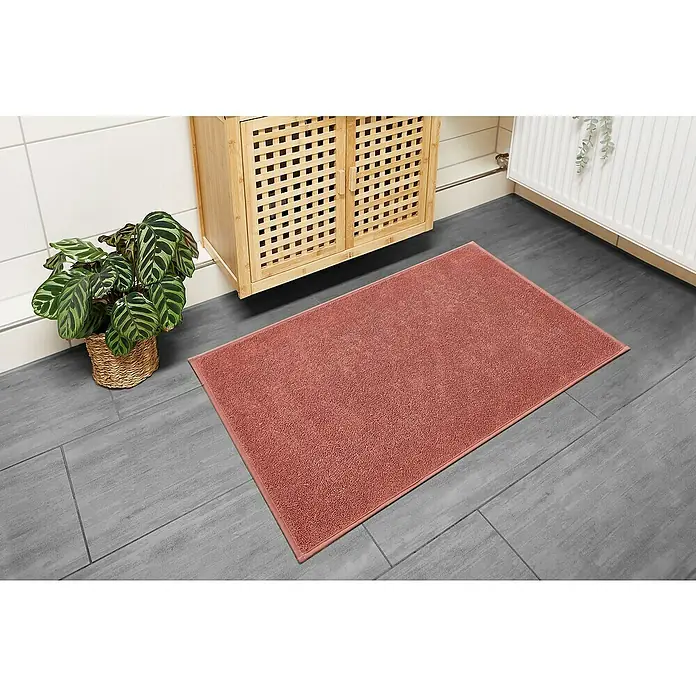 Badteppich Malibu - 60 x 100 cm, Rostrot, 100% Polyester - Nahaufnahme einer rosafarbenen Badematte auf grauem Fliesenboden, neben einem Holzschrank und einer Pflanze.