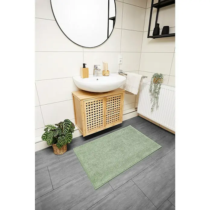 Badteppich Malibu - 60 x 100 cm, Salbei, 100% Polyester - Badezimmer mit mintgrünem Teppich vor einem Waschtisch aus Holz, ergänzt durch einen runden Spiegel und eine Regalablage.