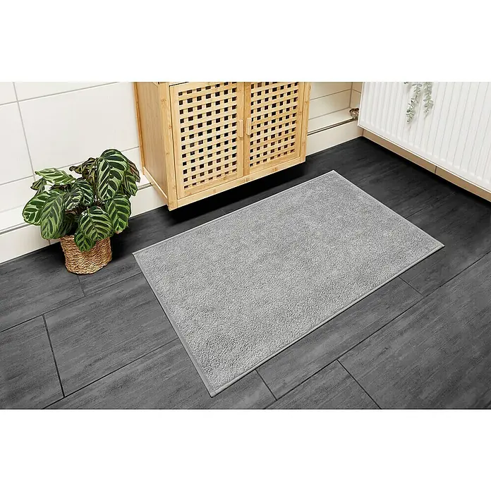 Badteppich Malibu - 60 x 100 cm, Grau, 100% Polyester - Graue Badematte liegt auf dunklen Fliesen neben einem Pflanzenkorb und einem Holzschrank, die Badezimmeratmosphäre ergänzend.