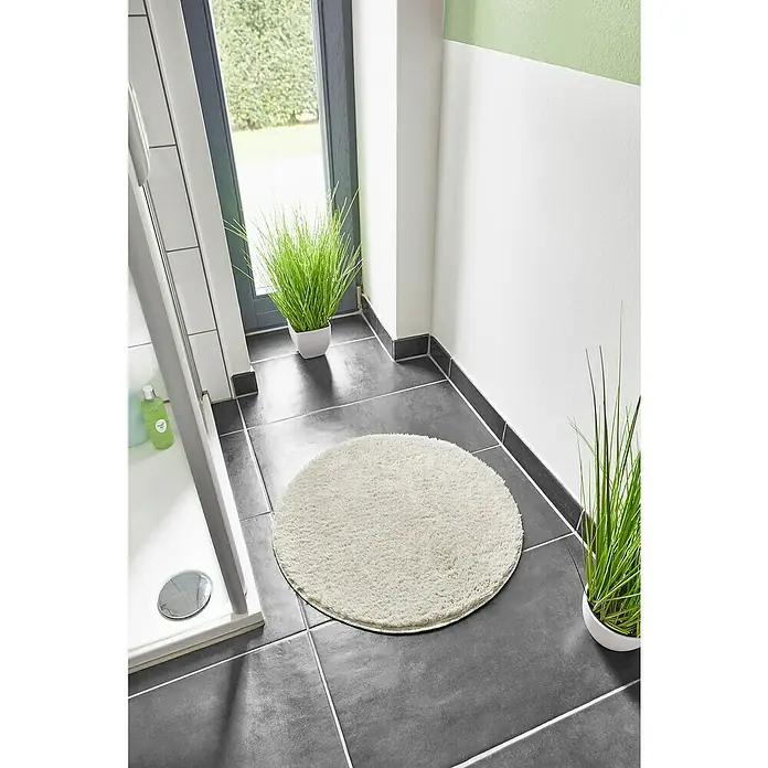 Badteppich Florida - 60 x 60 cm, Beige, 100% Polyester - Ein runder, weißer Badteppich liegt auf dunklen Fliesen im Badezimmer, neben einer Dusche und einer Grünpflanze.